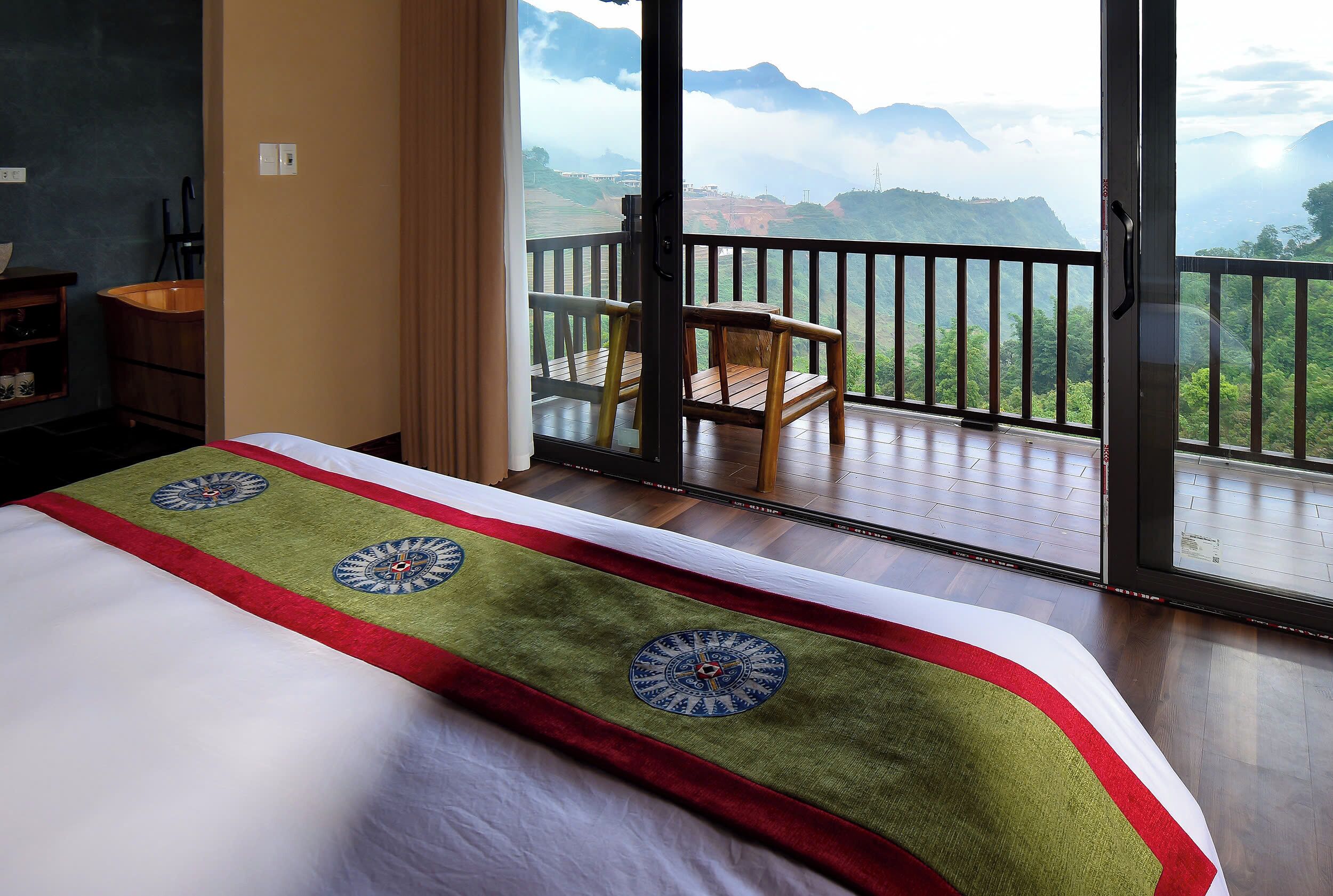 A hotel room with a bed, a wooden chair, and a balcony with a view of mountains.