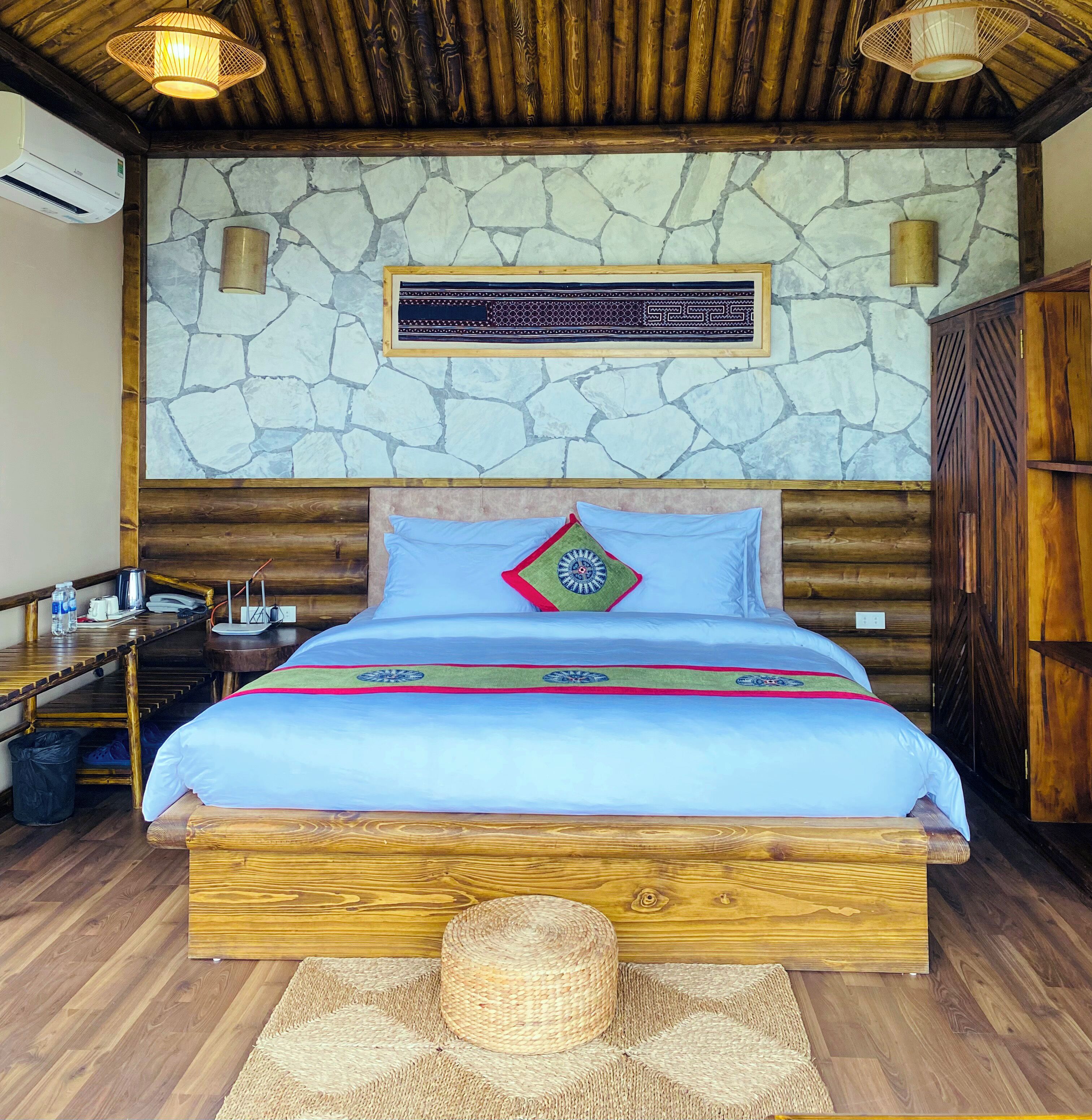 Una cama con estructura de madera, cubierta con un edredón azul y almohadas decorativas, una pared de piedra con un cartel enmarcado y un techo de madera con luces colgantes.