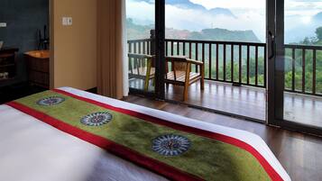 A hotel room with a bed, a wooden chair, and a balcony with a view of mountains.