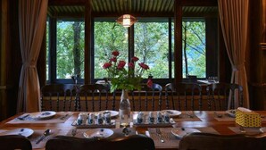 Une table à manger dressée pour six personnes, agrémentée d’un vase de fleurs rouges, de chaises en bois et offrant une vue sur les arbres à travers de grandes fenêtres.