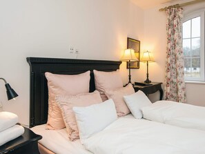 Ein ordentlich bezogenes Bett mit weißen Leinentüchern und mehreren Kissen, ein schwarzer Kopfteil und zwei Nachttischlampen mit floralem Vorhang.