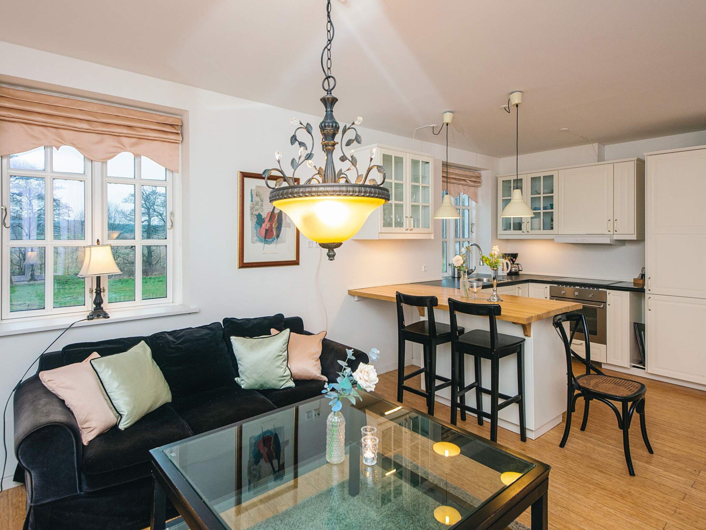 A modern kitchen with white cabinets, a dining area with a glass table, and a black sofa with cushions.