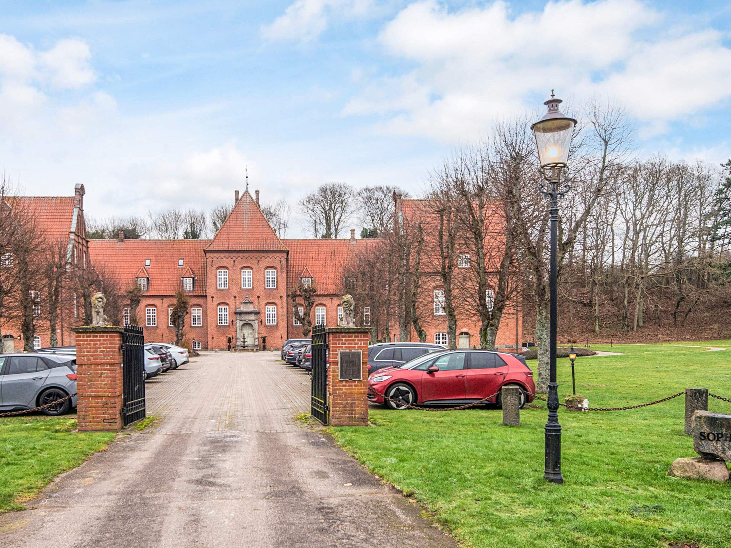 A red brick building with a central tower, a paved driveway leading to a gated entrance, and a parking area with cars.