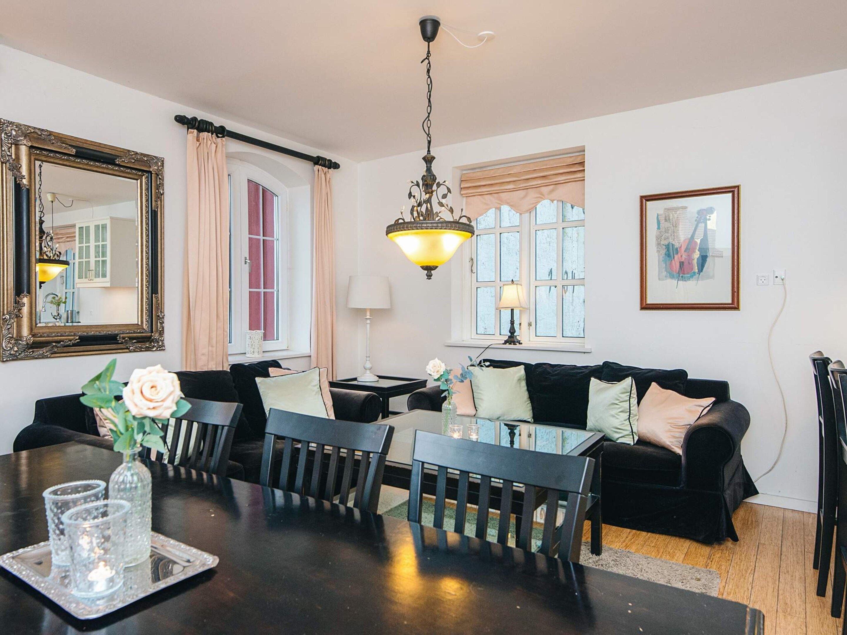 A dining area with a table, chairs, and a chandelier. There is a sofa and a window with curtains.