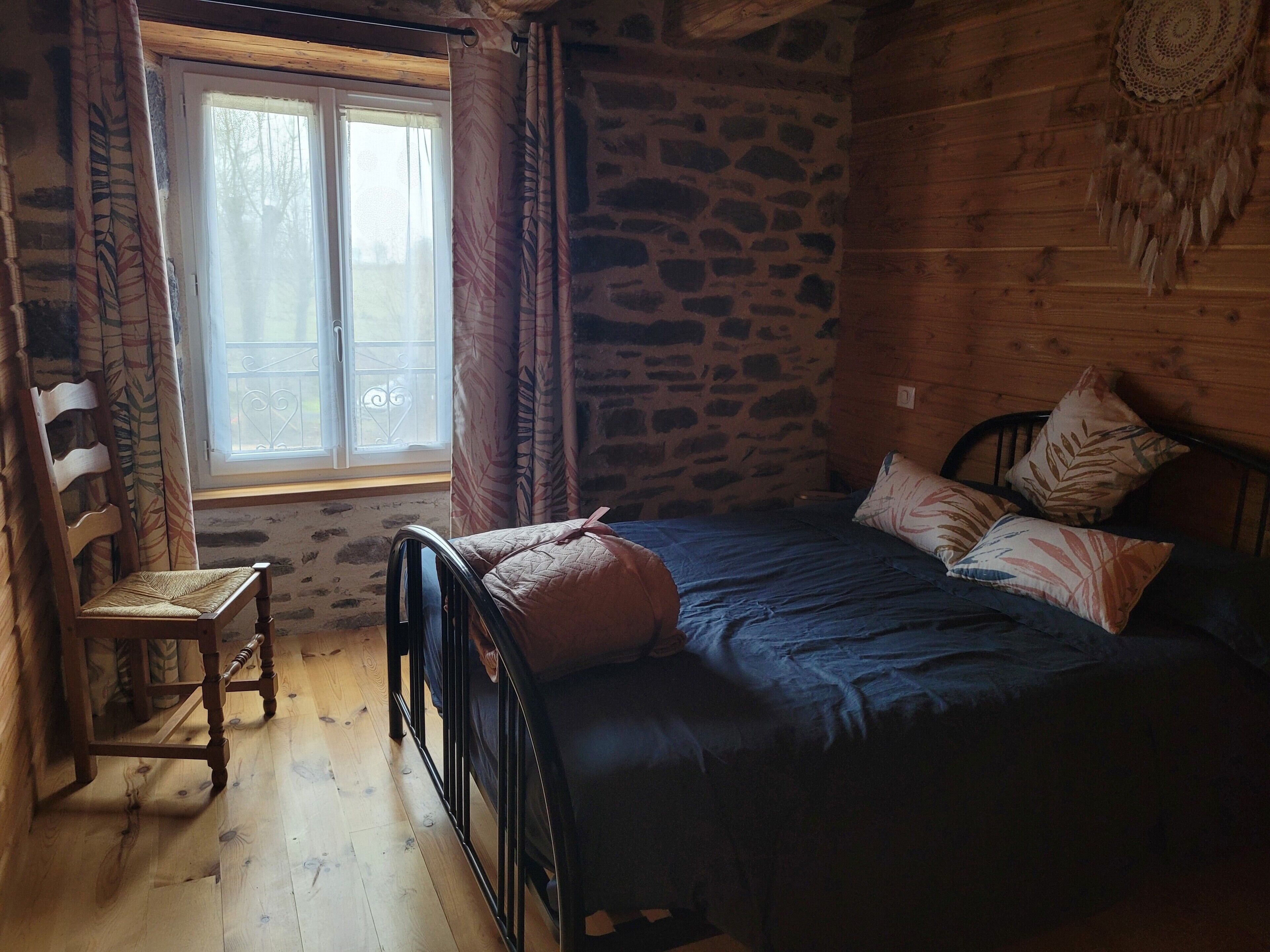 Une pièce de cabane en bois comprenant un lit, une chaise, une fenêtre avec des rideaux et un mur en pierre.