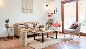 A living room with a beige sofa, a coffee table, a side table with a plant, a chair with a cushion, and a large window with curtains. - Killay A4 - Villa de lujo de 2d (Cusco)
