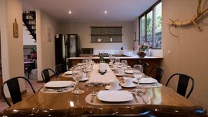 Un comedor con una mesa larga dispuesta para una comida, una cocina al fondo y una escalera que sube a otro nivel.