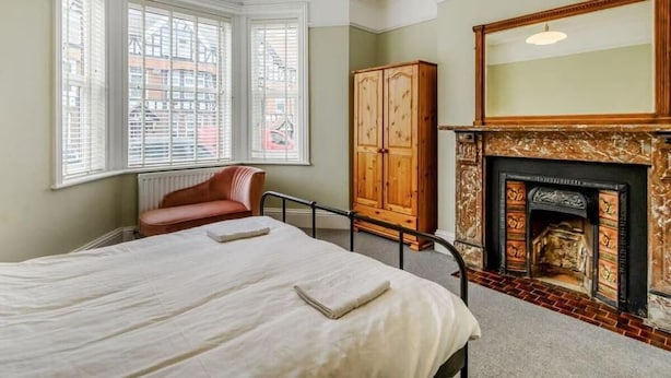 A bedroom with a large bed, a wooden wardrobe, a fireplace, and a window with blinds. - Ground Floor Apartment Beverley House (Cromer)