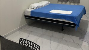 Una habitación pequeña con cama individual, una colcha azul, almohadas blancas, una cortina gris y un equipo de aire acondicionado blanco en la pared.