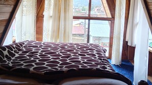 A room with a bed, wooden walls, a window with curtains, and a view of buildings and mountains.