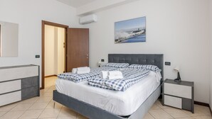 Un dormitorio con una cama, mesitas de noche, aire acondicionado montado en la pared, una puerta y un cuadro enmarcado.