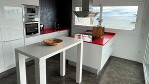 Una cocina moderna con armarios blancos, encimera roja, horno empotrado y ventana con vistas al mar.