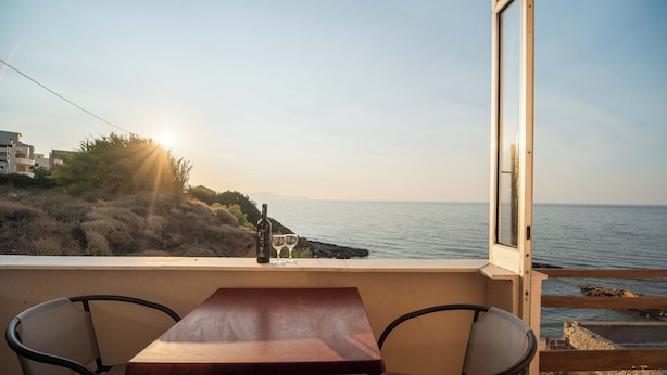 A balcony with a wooden table and two chairs, overlooking a coastal landscape with the sea and hills. - SEA GEM APARTMENTS steps from the sand (Chania)