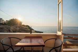Un balcón con mesa de madera y dos sillas, con vista a un paisaje costero que incluye el mar y colinas.