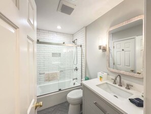 Un baño moderno con ducha de vidrio, inodoro blanco, lavamanos con espejo y un perchero para toallas.