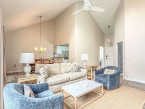 Ein geräumiges Wohnzimmer mit hoher Decke, einem Deckenventilator, einer Sofagarnitur, zwei Sesseln, einem Couchtisch und einer im Hintergrund sichtbaren Küche.