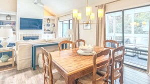 A modern living room with a wooden dining table, comfortable seating, a TV, and a large window with a view. - Quiet Sea Pines Home: Screened Porch + Beach Gear (Hilton Head Island)