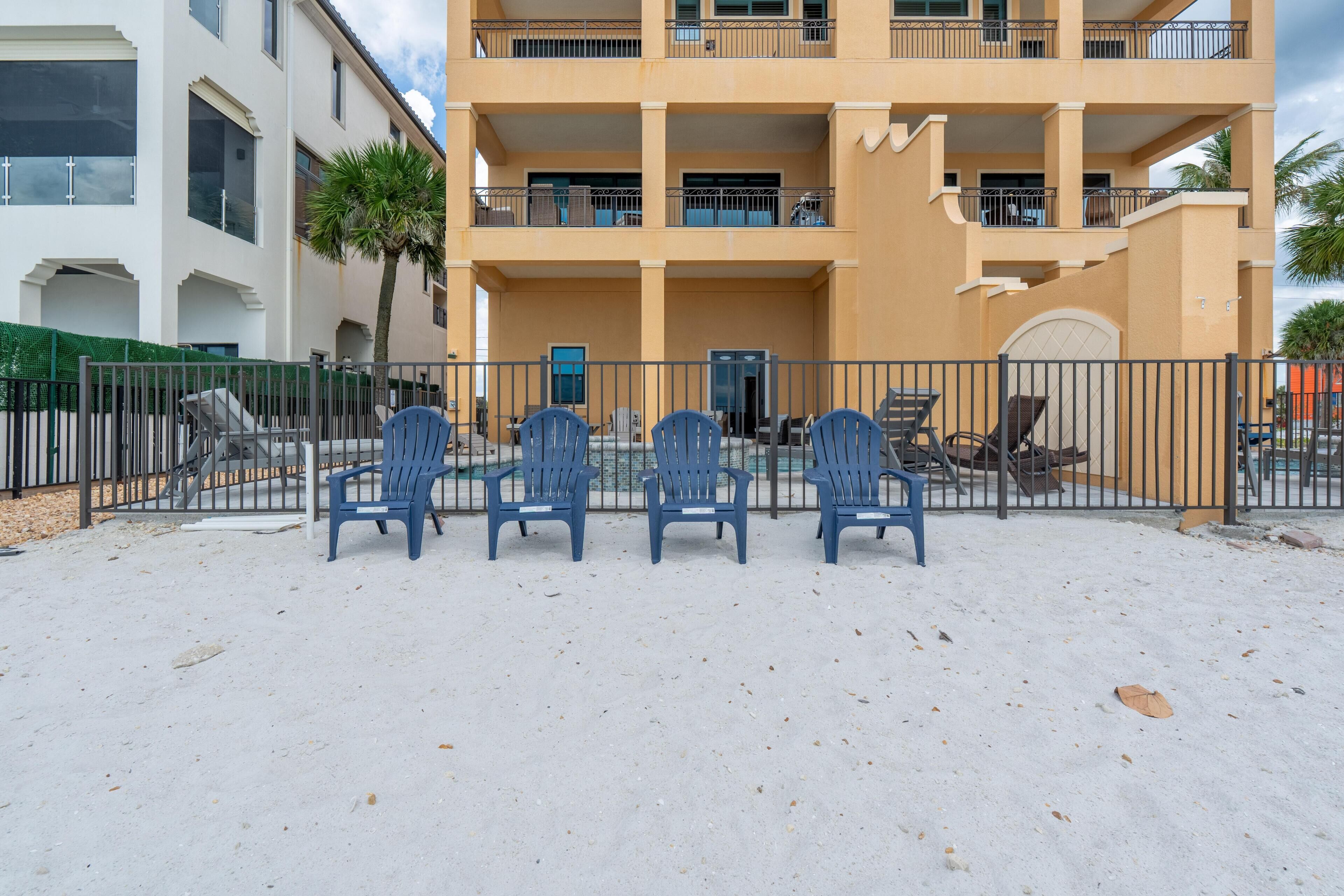 A multi-story building with a balcony, outdoor seating area with chairs, and a sandy ground.