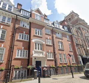 A row of red-brick townhouses with white window frames, a central entrance with a sign, and a black metal fence. - Incredibly Located 1BDR Hideaway in Soho (Greater London)