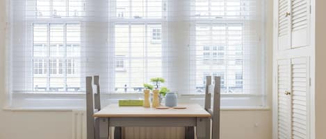 A room with a table and chairs near a window with blinds, a radiator below, and a door with louvered shutters.