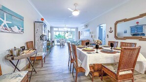 A spacious dining area with a long table set for six, surrounded by wicker chairs. A ceiling fan is visible, and the room has a nautical theme with starfish decorations and a large window offering a view of the outdoors. - Apartamento Enseada 24 (Guarujá)
