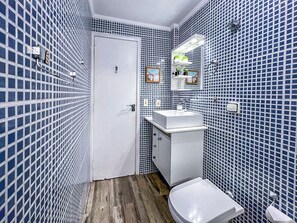 Kylpyhuoneessa on valkoinen pesuallas, peili ja wc-istuin. Seinät on päällystetty sinisillä mosaiikkilaatoilla.