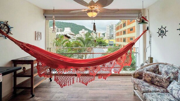 A room with a hammock, a ceiling fan, a sofa, and a view of buildings and greenery. - Apartamento Enseada 24 (Guarujá)