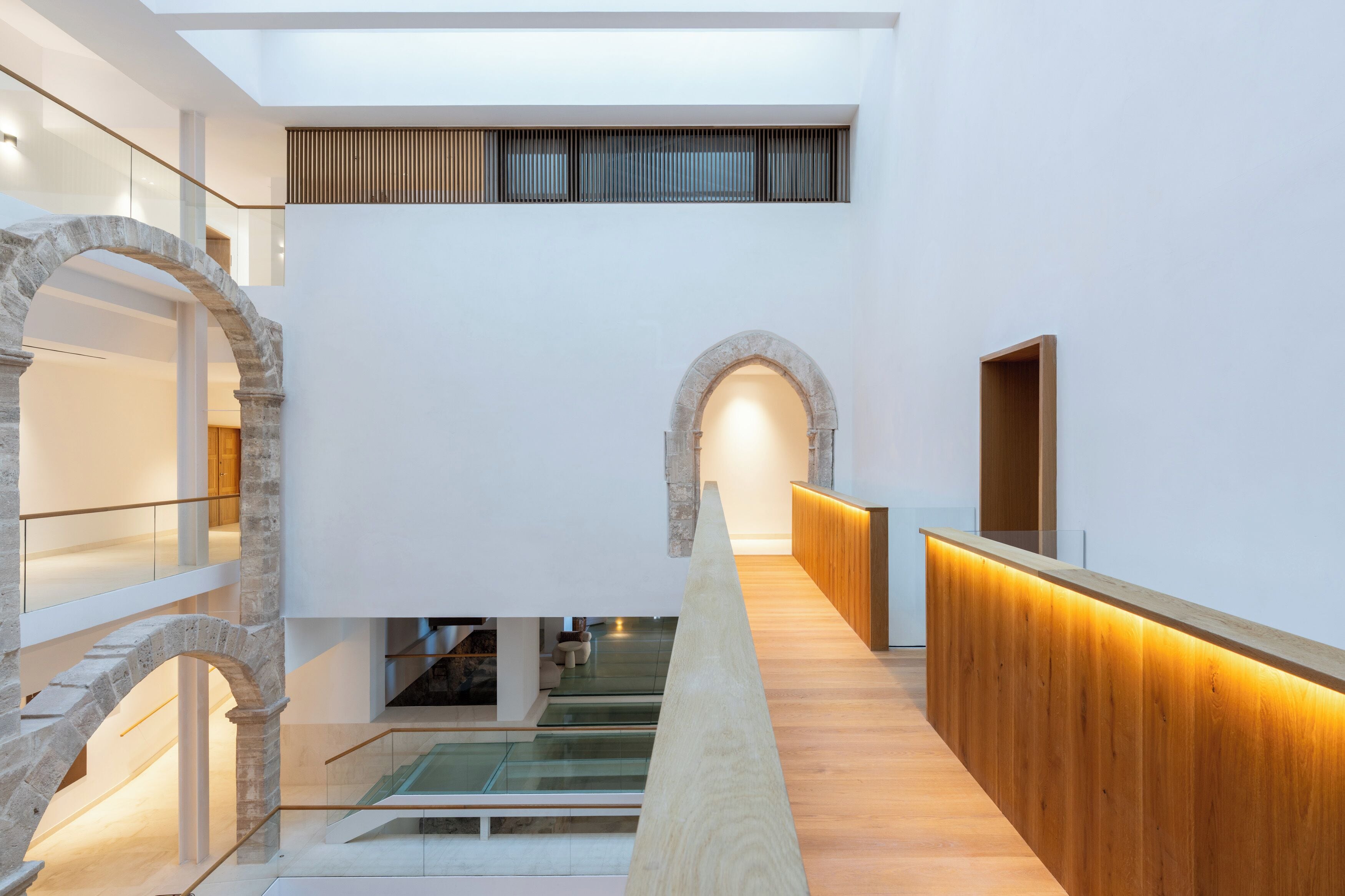 Un intérieur moderne avec un comptoir d’accueil central en bois, des arcades en pierre et un escalier en verre.