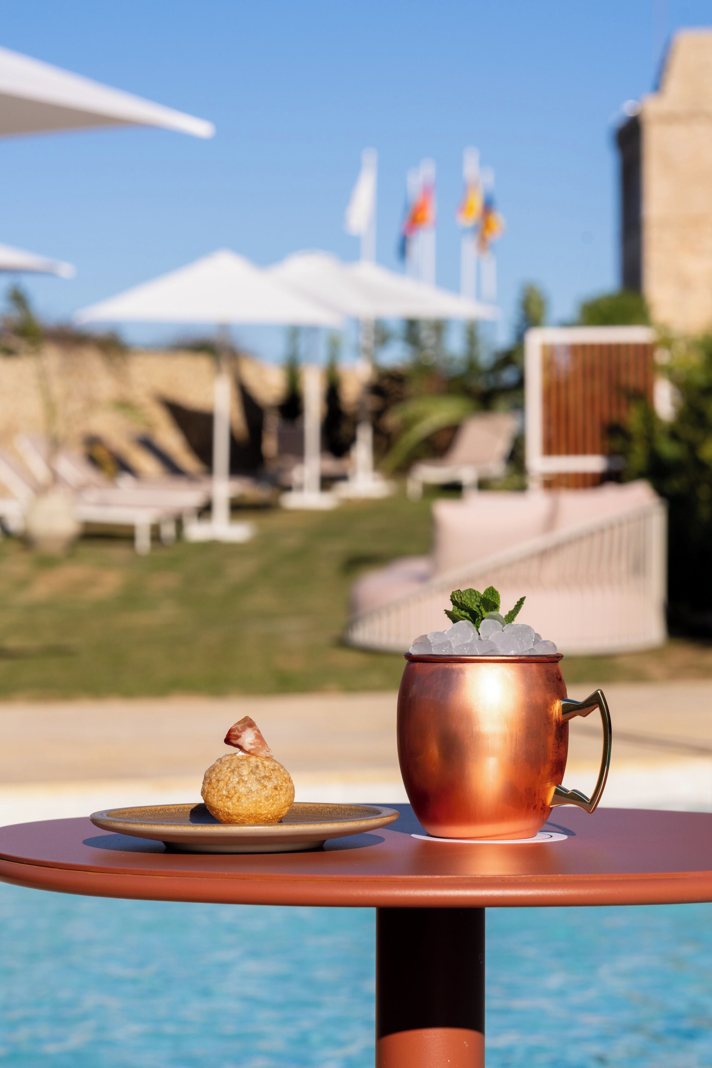 Una taza de cobre con hielo y una galleta sobre una mesa junto a una piscina.