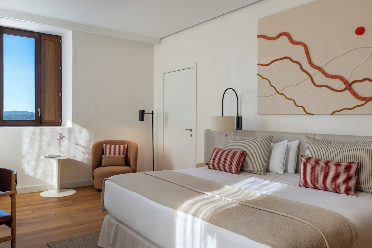 Une chambre d’hôtel avec un grand lit, une chaise, une petite table avec un vase et un tableau au mur.