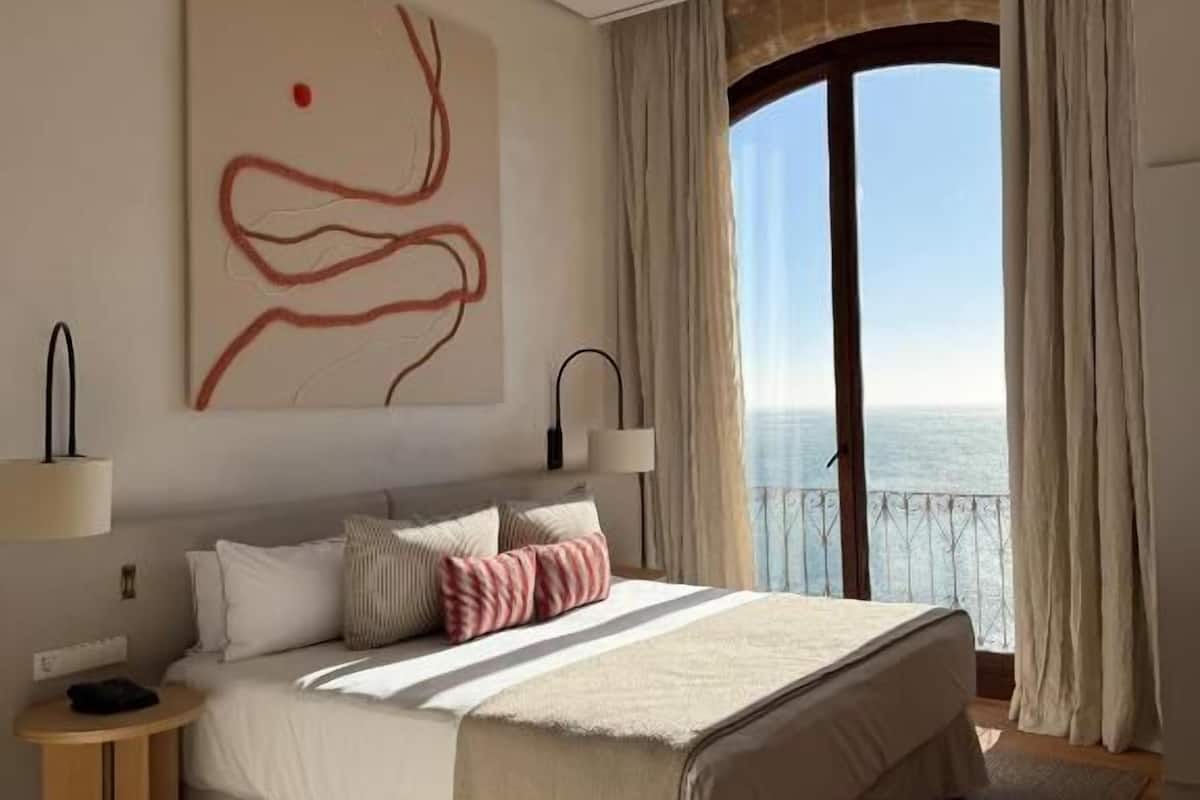 Une chambre avec un grand lit, une vue sur la mer et une peinture abstraite moderne accrochée au mur.