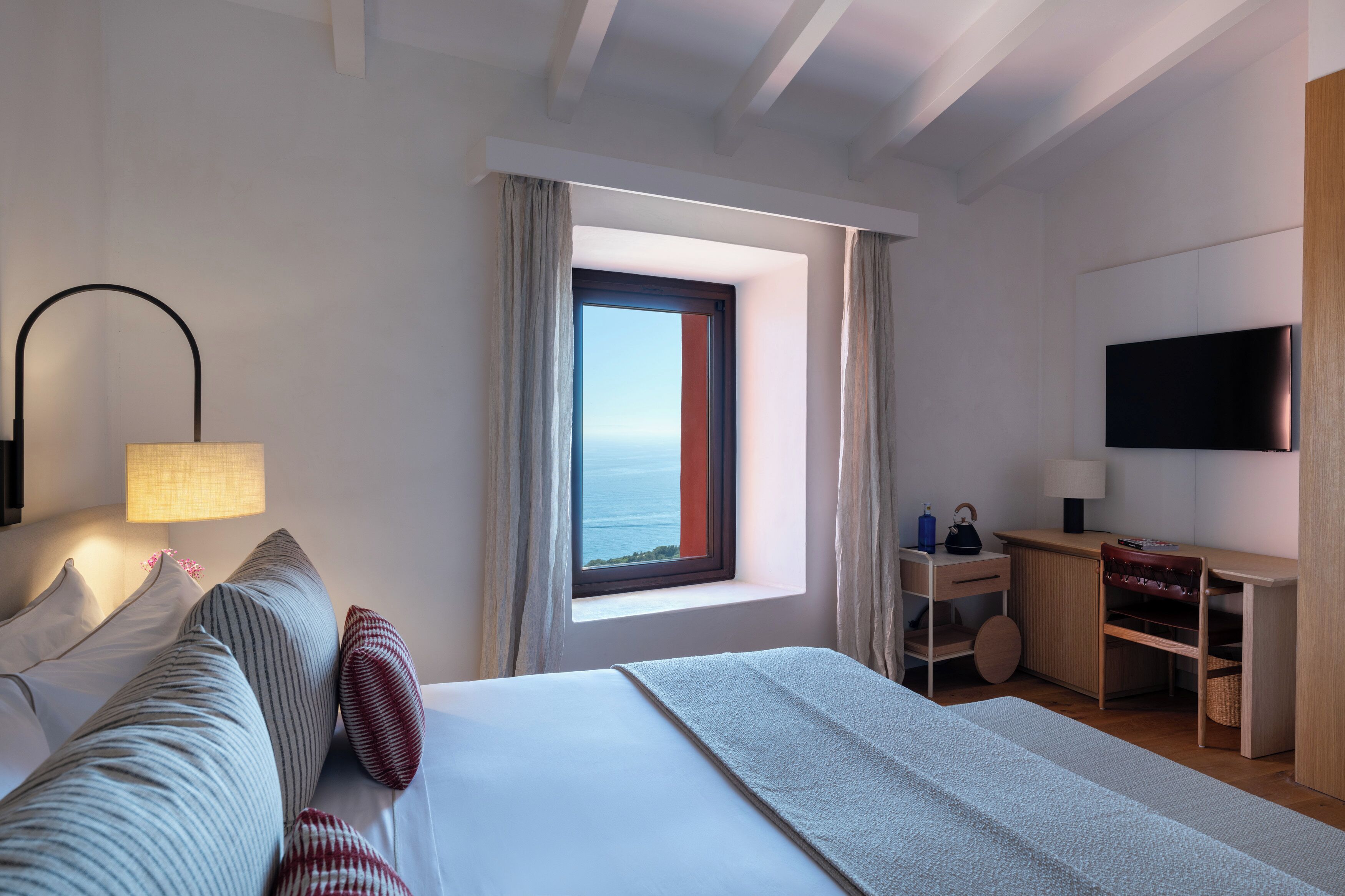 A hotel room with a bed, a desk, a TV, and a window with a view of the sea.