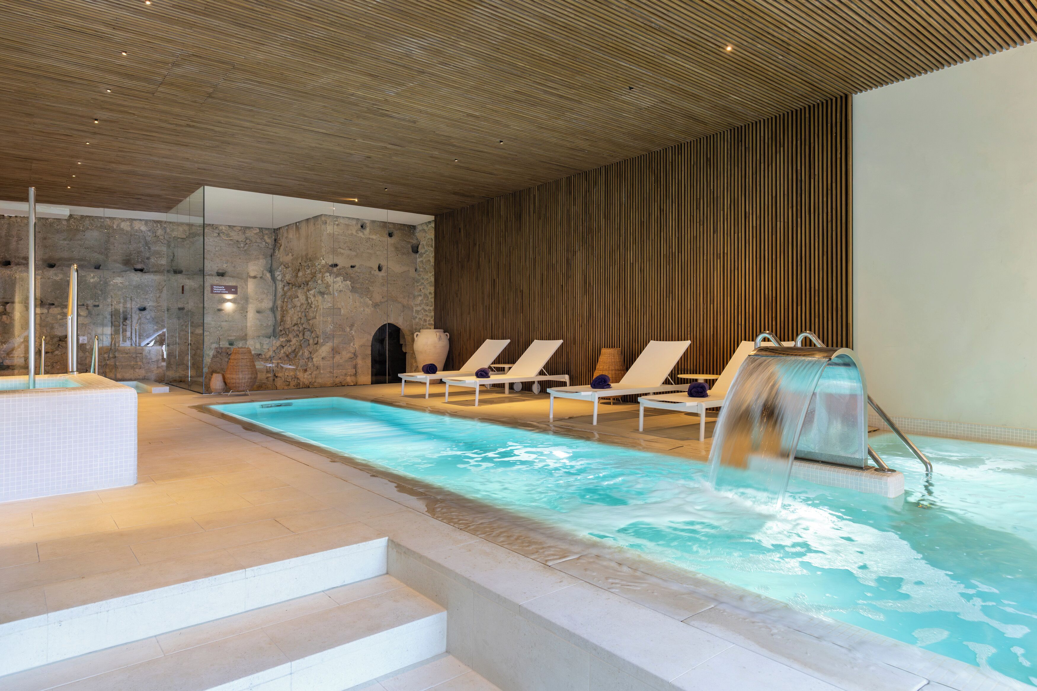 A modern indoor pool with clear water, white lounge chairs, a stone wall, and a wooden ceiling.