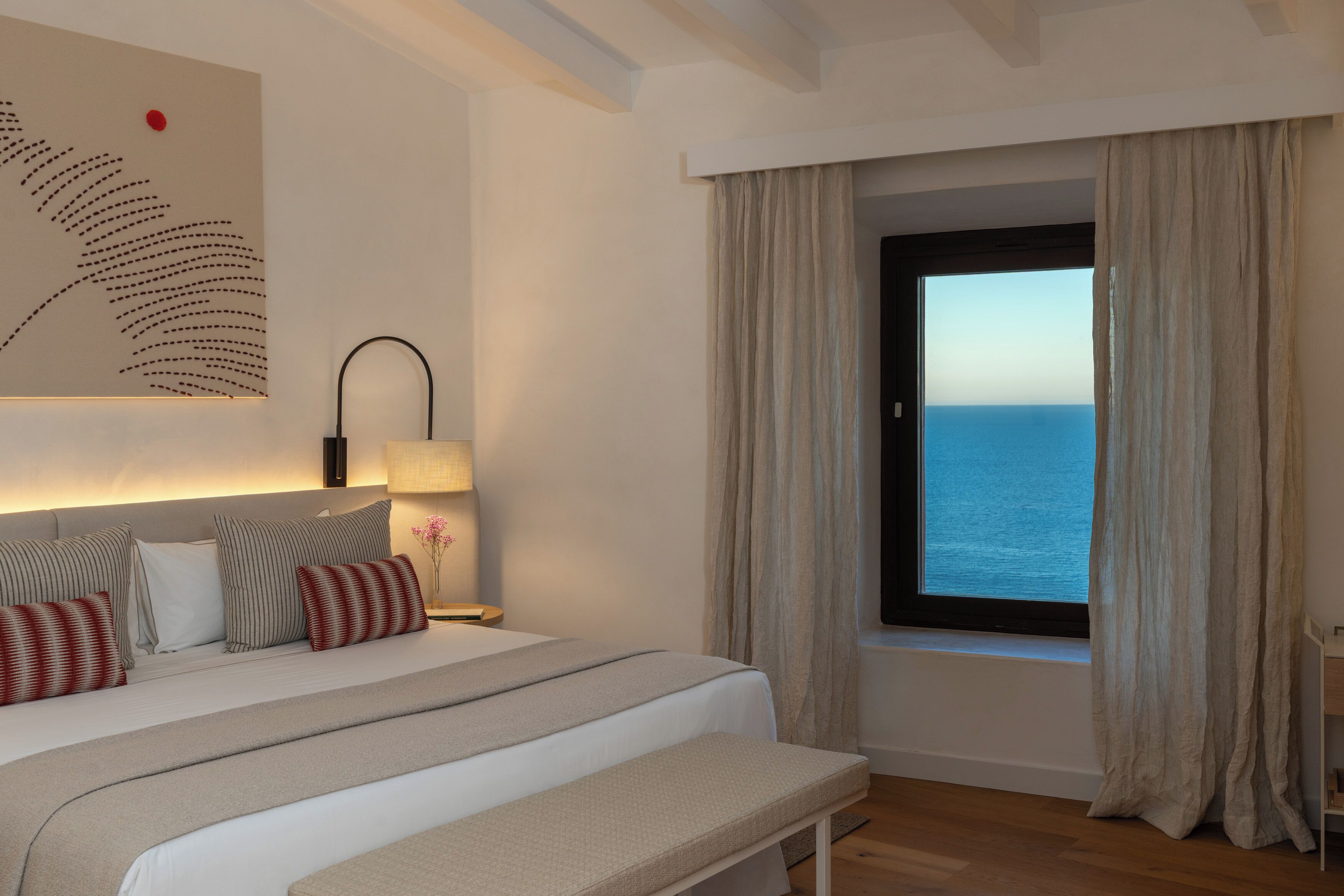 A bedroom with a large bed, a bedside table with a lamp, a view of the sea through a window with curtains, and a wall art piece.