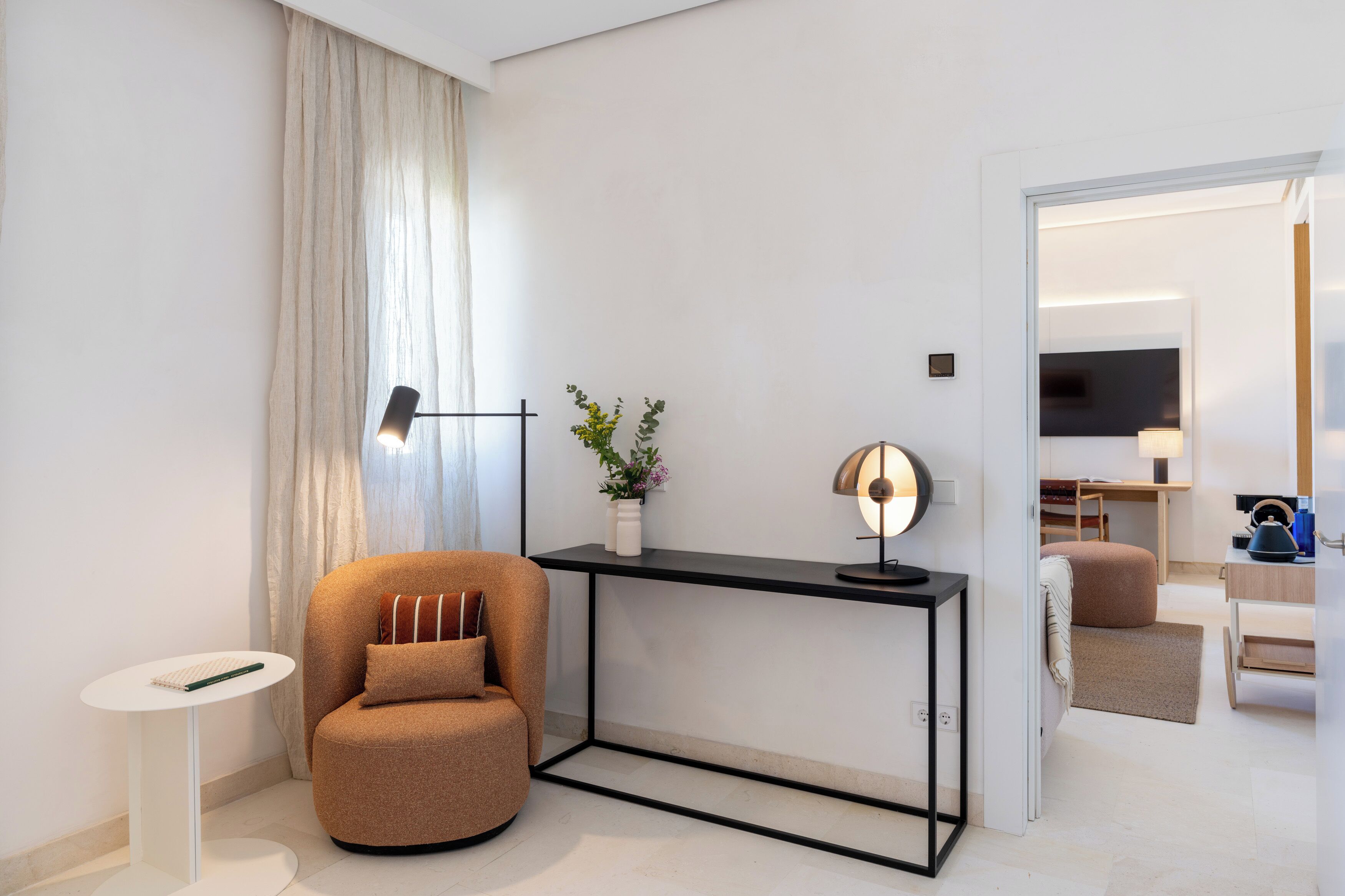 Quarto moderno com poltrona arredondada, pequena mesa branca, mesa consola preta com uma luminária e uma planta, e uma TV ao fundo.