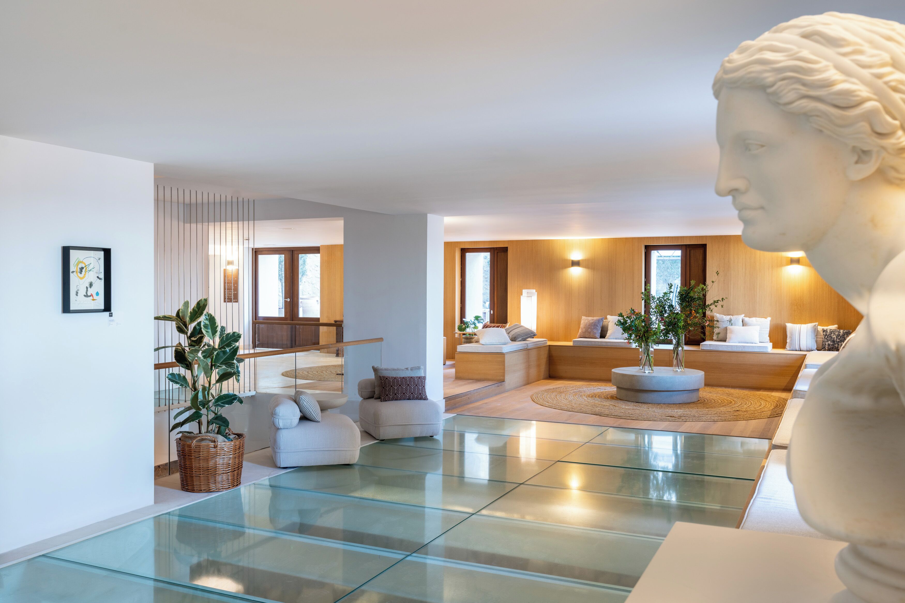 Interior moderno com piso de vidro, sofá branco, mesa de centro redonda e uma escultura de busto.
