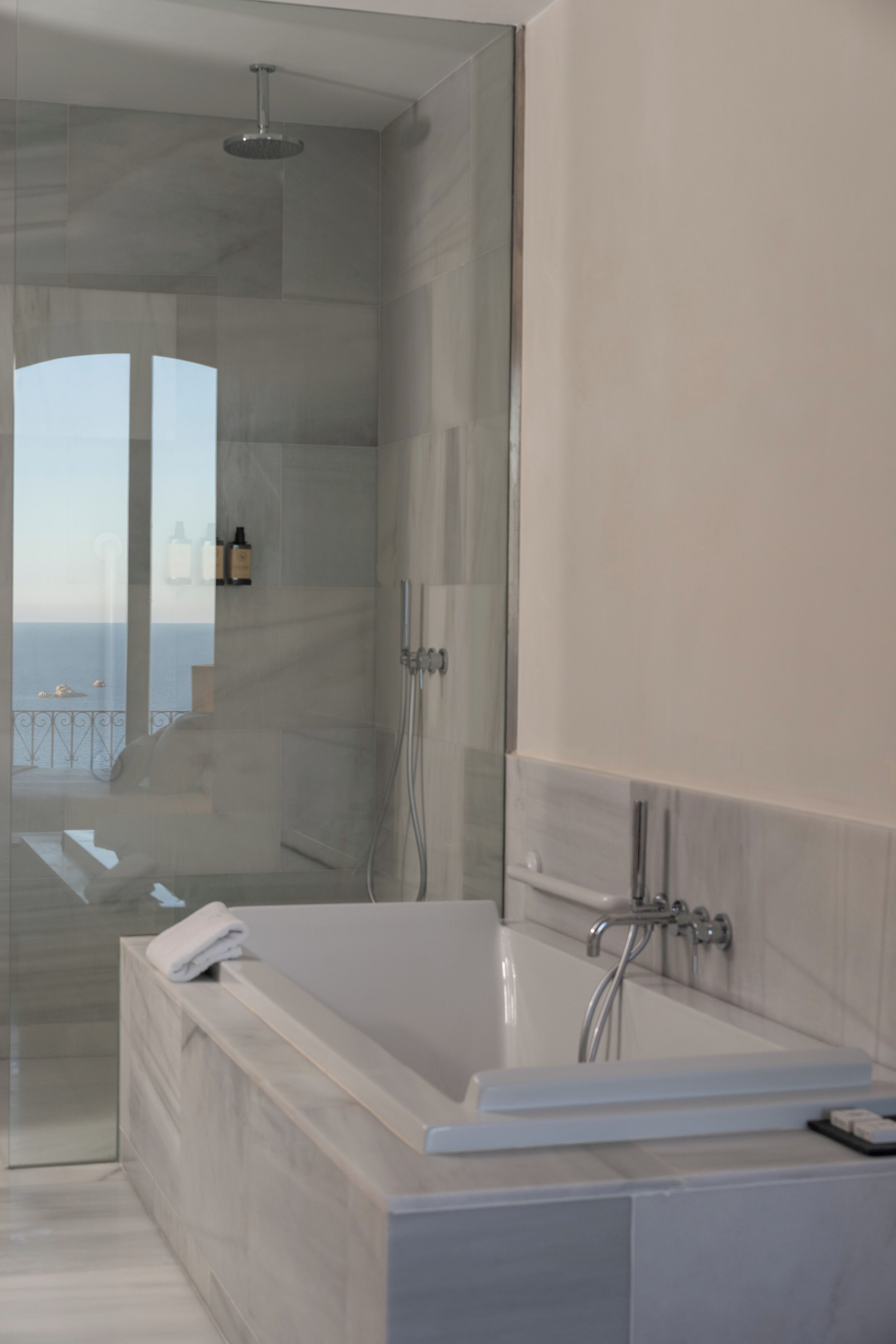 A modern bathroom with a glass-enclosed shower, a large bathtub, and a view of the sea.