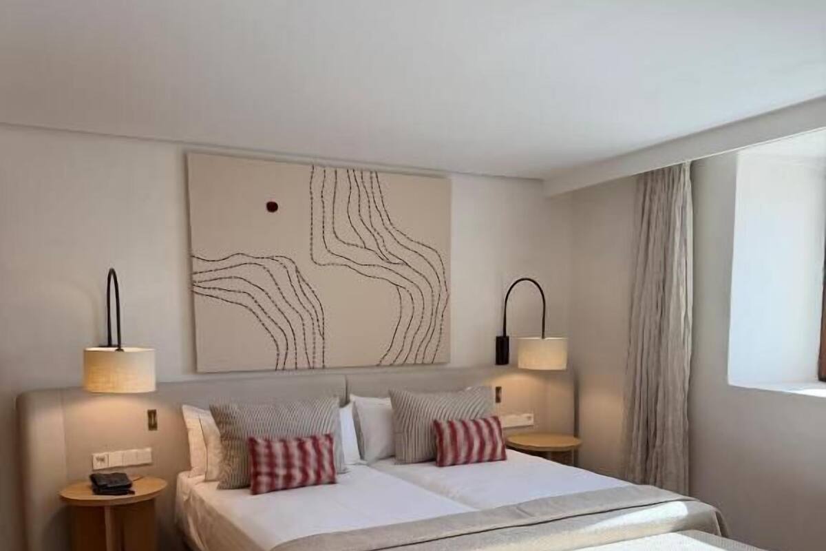 Une chambre moderne avec un lit double, des lampes de chevet, un banc et une grande œuvre d’art abstraite accrochée au mur.