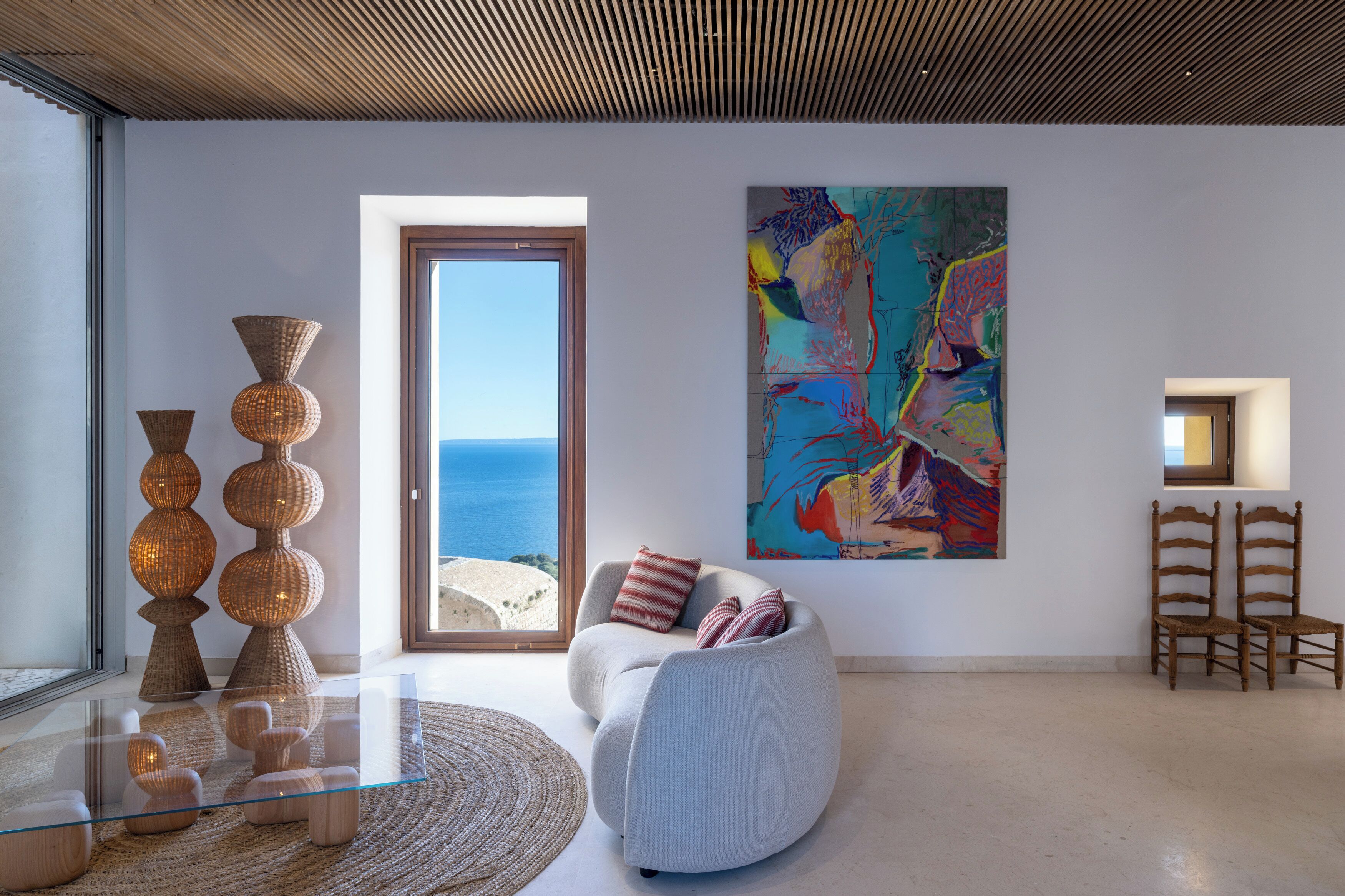 A modern living room with a large window, a colorful abstract painting, and a glass coffee table.