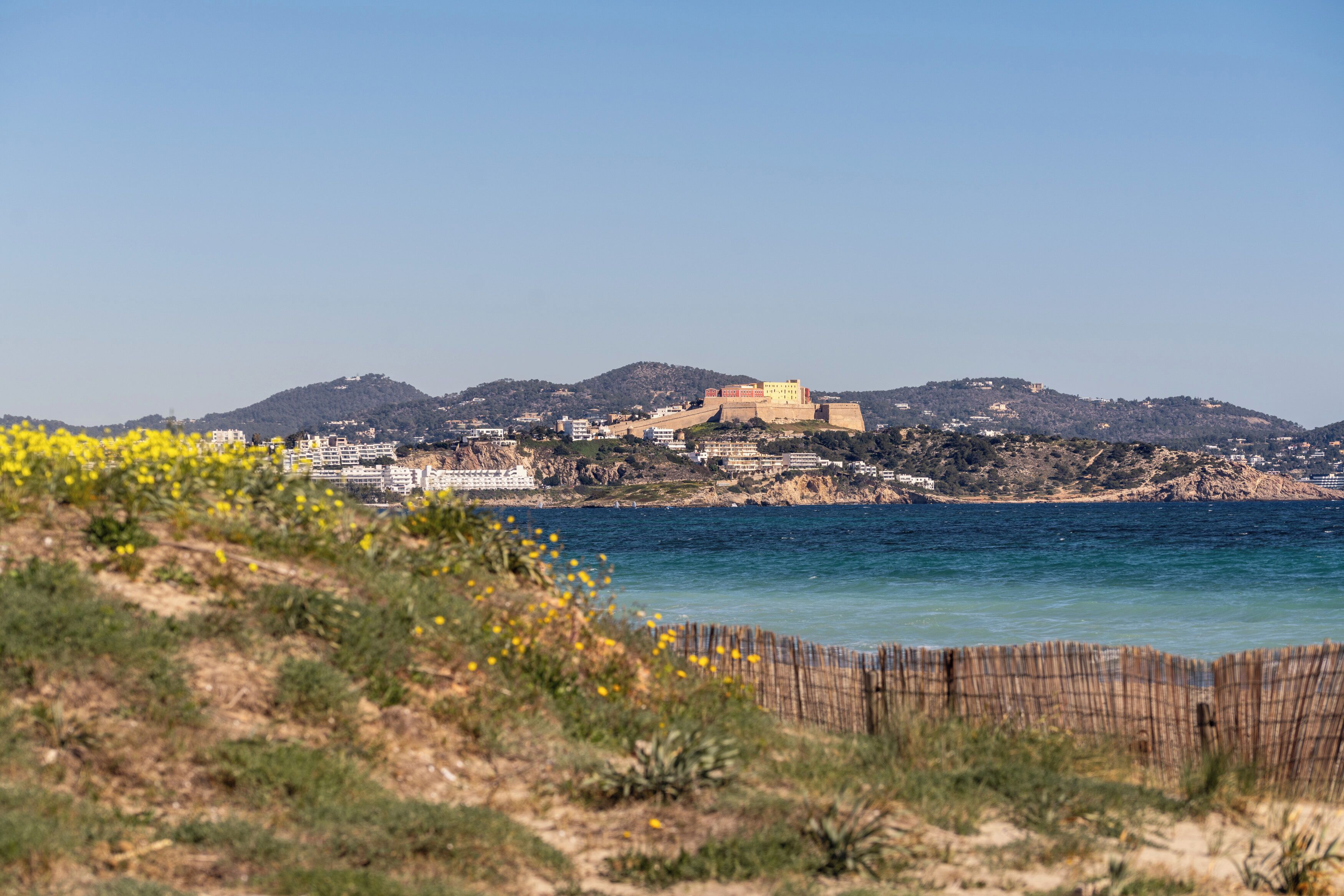 Vista costera con un fuerte histórico en una colina, que domina una playa y un pueblo.
