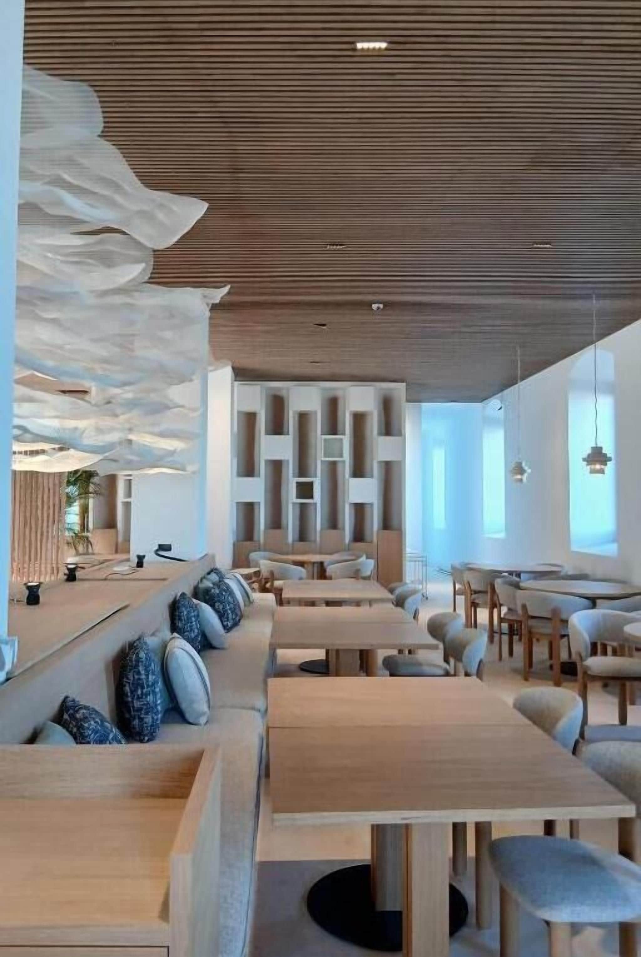 A modern restaurant interior with wooden tables and benches, a ceiling with a cloud-like design, and a bright, airy atmosphere.