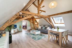 Quarto estilo loft com vigas de madeira aparentes, área de refeições com mesa e cadeiras, um sofá e vista por um claraboia.