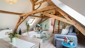 Habitación estilo loft con vigas de madera a la vista, zona de comedor con mesa y sillas, sofá, televisor y escalera.
