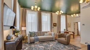 Un salón con sofá, sillón y alfombra estampada. Hay un televisor a la izquierda, una lámpara y una planta sobre la mesita de noche.