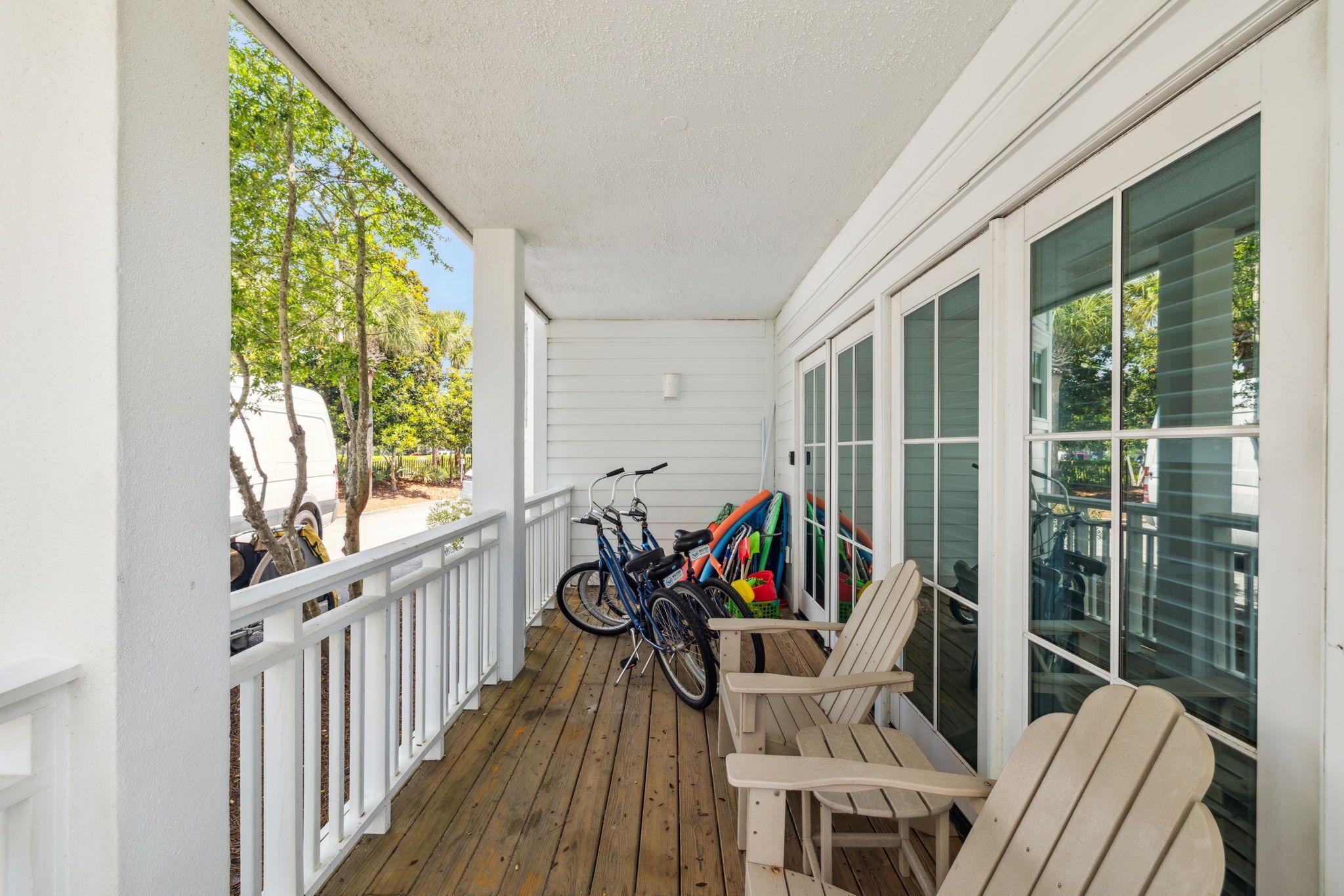 A covered porch with a wooden floor, white railings, and large windows. There are bicycles and colorful children's play equipment.