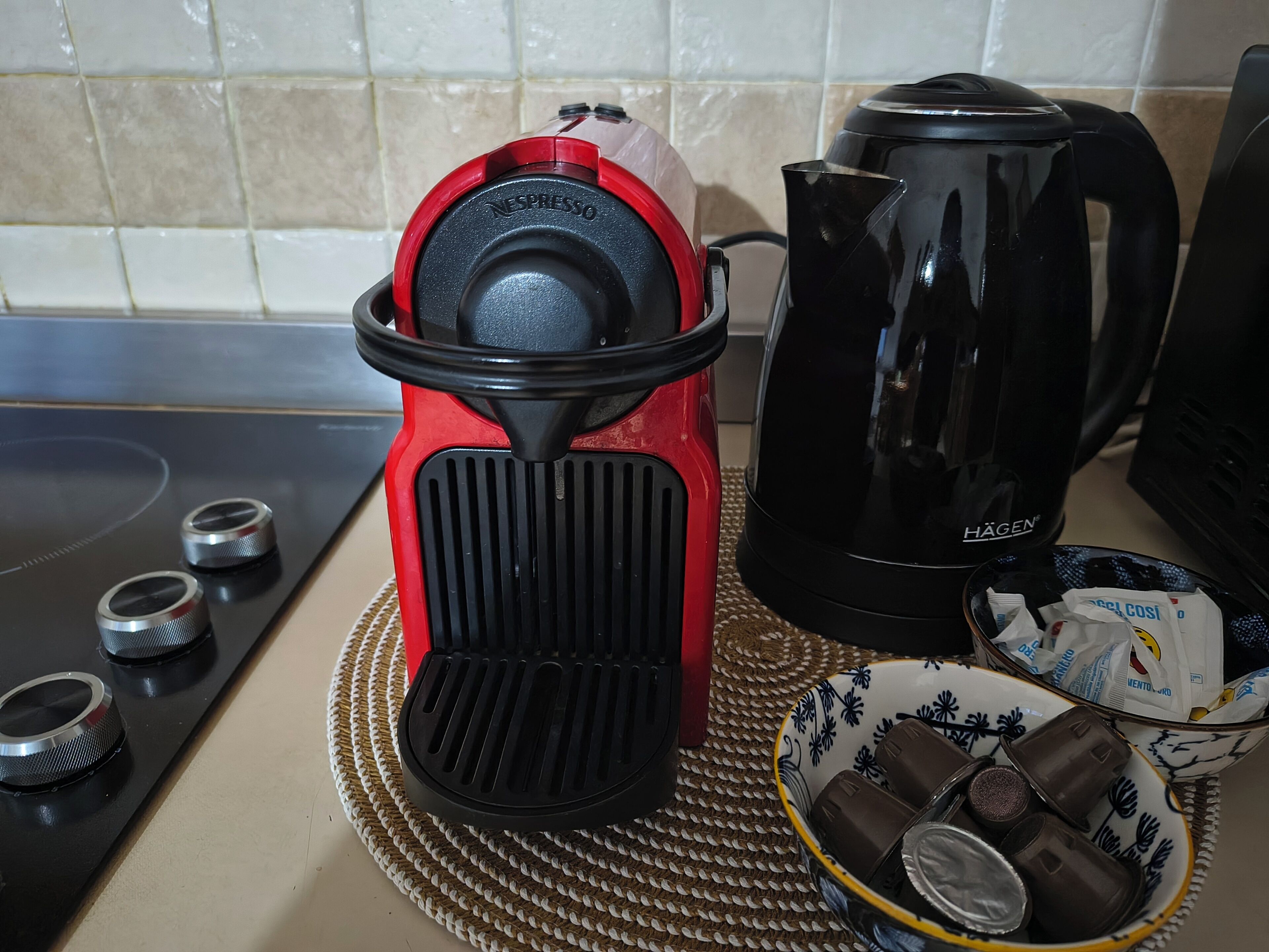 Une cuisine avec une machine à café rouge et noire, une cafetière noire et un bol de dosettes de café posé sur un set de table en fibres tressées.