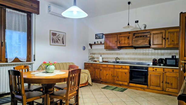 Une cuisine avec des meubles de rangement en bois, une table à manger avec des chaises, un luminaire suspendu et une fenêtre avec un rideau.