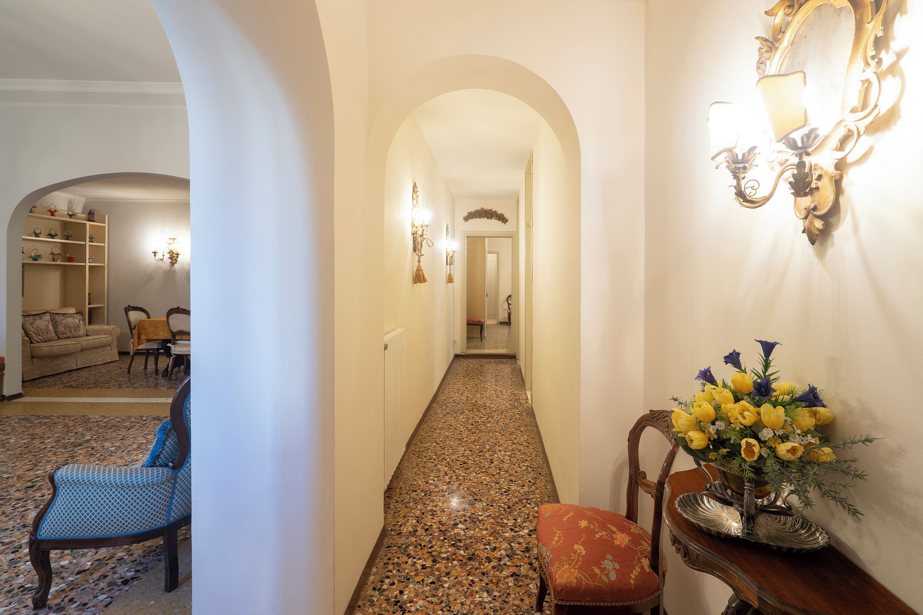 Un corridoio con soffitto a volta, una sedia, un tavolo con un vaso di fiori e una mensola con diversi oggetti.