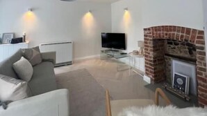 A modern living room with a sofa, a glass coffee table, a flat-screen TV, a fireplace, and a ceiling light fixture. - Samphire House (Cromer)