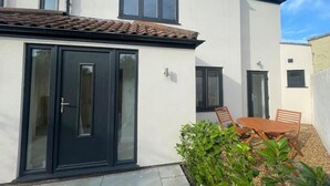 A modern two-story house with a tiled roof, a black front door, and a small patio area with a wooden table and chairs. - Samphire House (Cromer)